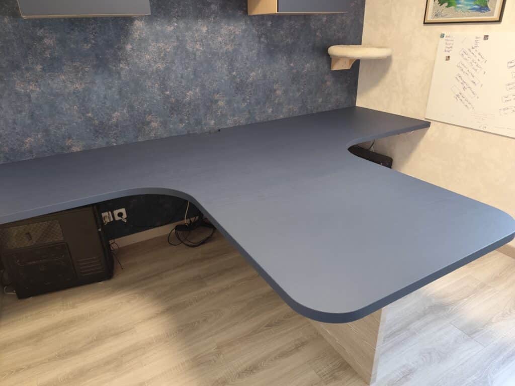 Bureau d'angle en L bleu-gris avec tour de PC, câbles. Mur texturé bleu, niche de chat moelleuse et tableau blanc notes "Noeline". Sol parquet clair.