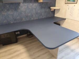 Bureau d'angle en L bleu-gris avec tour de PC, câbles. Mur texturé bleu, niche de chat moelleuse et tableau blanc notes "Noeline". Sol parquet clair.