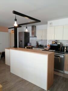 Cuisine ouverte moderne avec îlot central blanc et bois clair, plan de travail sombre, meubles crème, et luminaires suspendus noirs au plafond.