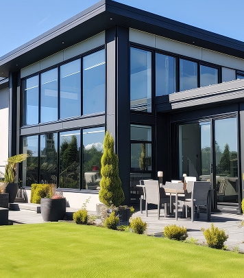 Maison moderne à grandes baies vitrées noires sur deux étages, pelouse verte bien entretenue et terrasse avec mobilier de jardin. Jour ensoleillé.