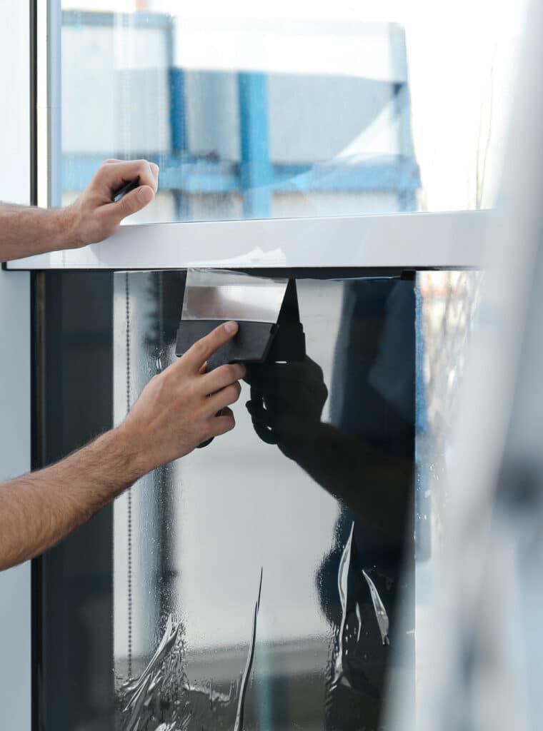 Pose film teinté vitre : Protection solaire et intimité Deux mains posent et lissent un film de protection noir sur une vitre avec une raclette. Des bulles et de l'eau sont visibles.
