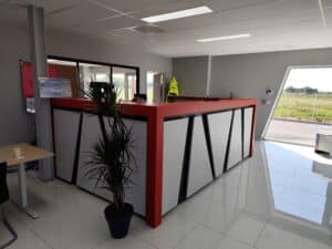 Comptoir de réception moderne rouge/blanc, sol brillant. Panneau "Retirez votre commande e-shop". Grande baie vitrée, plante. Produits CAIROX.