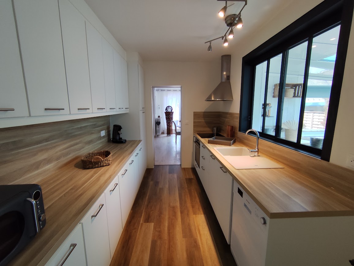 Cuisine moderne avec meubles blancs, plan de travail en bois, évier blanc et verrière intérieure noire.